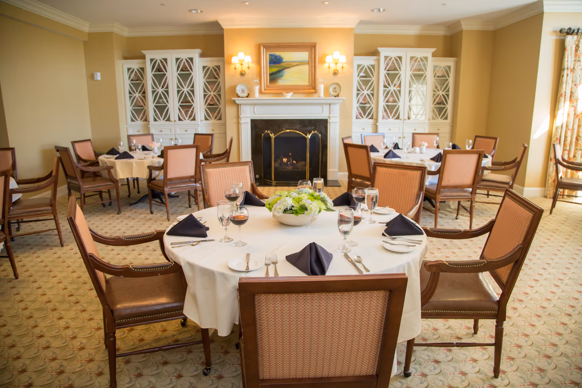 A warmly lit dining room with round tables covered in white tablecloths, set with plates, glasses, silverware, and black folded napkins. The room features wooden chairs with cushioned seats, a fireplace with a painting above it, and built-in cabinets with glass doors on either side of the fireplace.