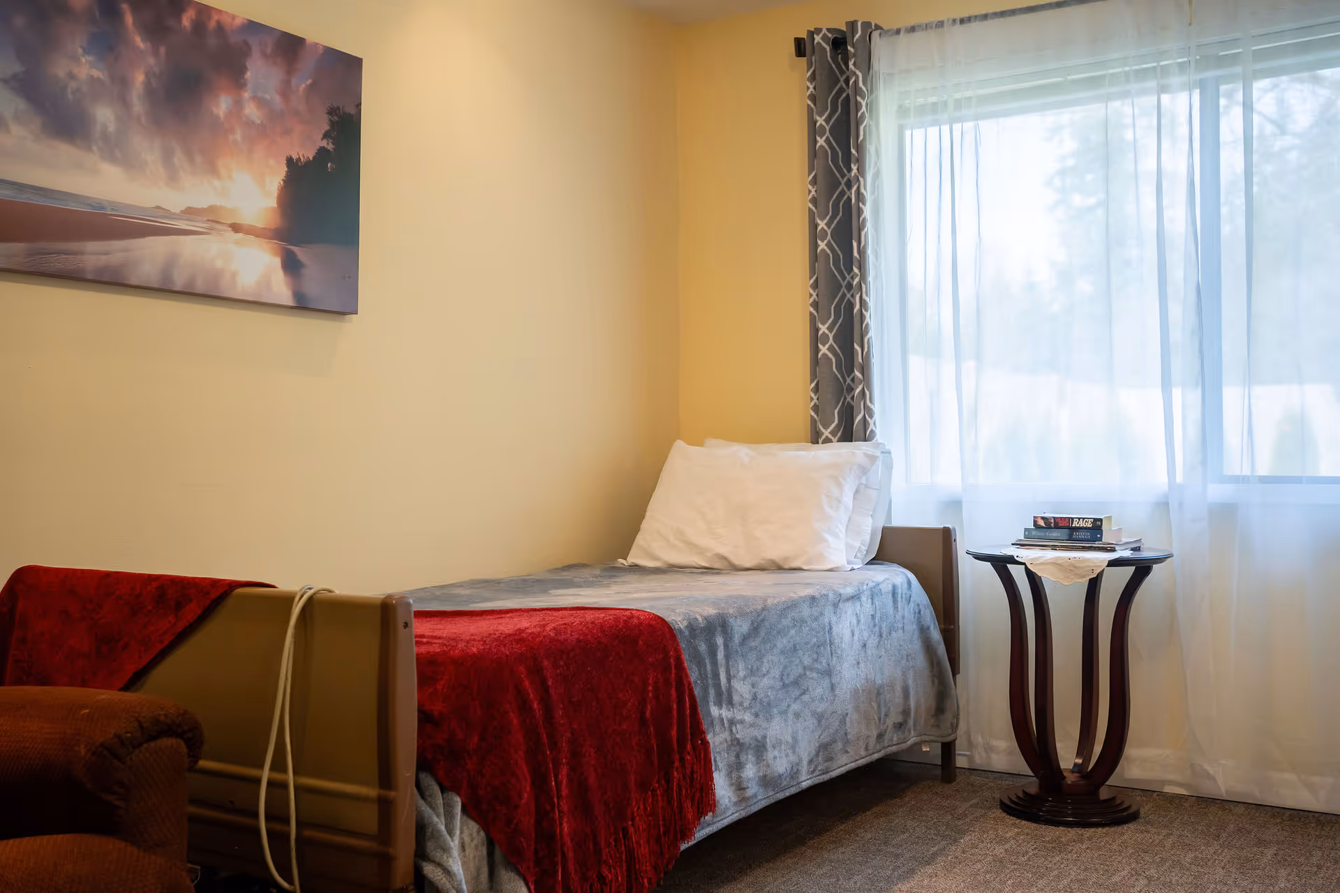 A small bedroom with a single bed covered with a gray blanket and a red throw. A white pillow is placed at the head of the bed. Next to the bed is a round wooden side table with a few books on it. The room has a large window with sheer white curtains and patterned drapes. A landscape painting hangs on the wall above the bed.