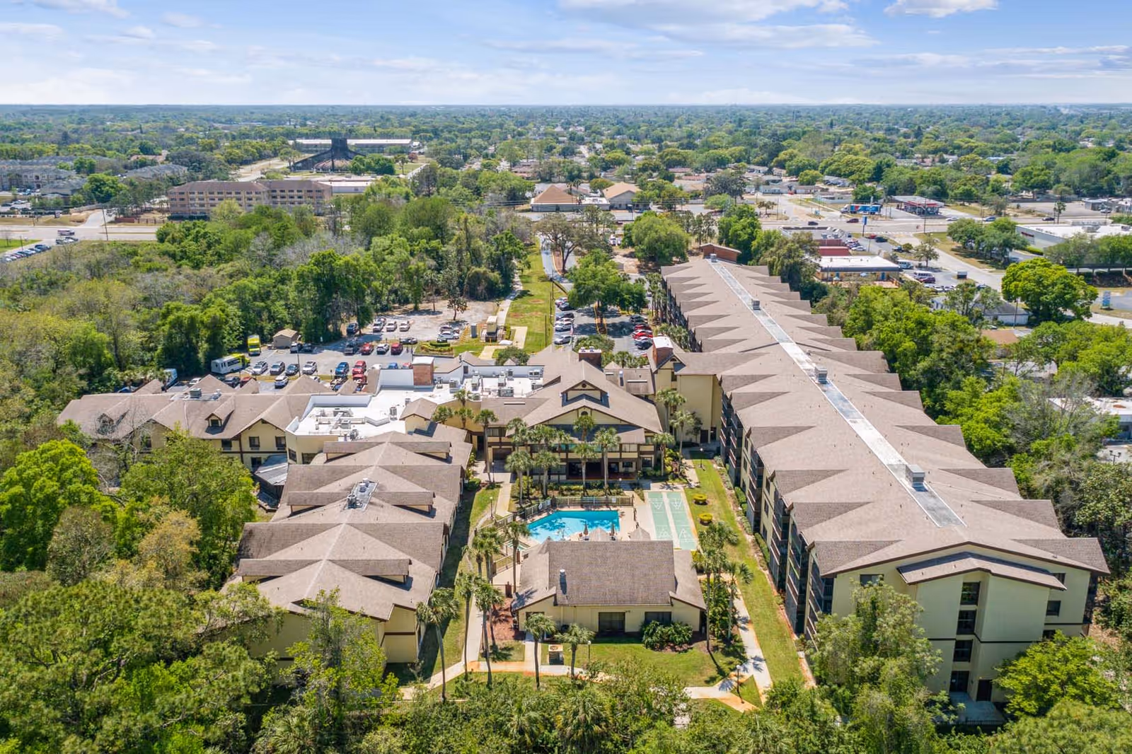 Aerial view of Vitality Living Baypoint Village senior living facility showing multiple connected buildings with beige roofs, a central swimming pool, shuffleboard courts, surrounding greenery, parking lots, and a suburban neighborhood in the background under a partly cloudy sky.