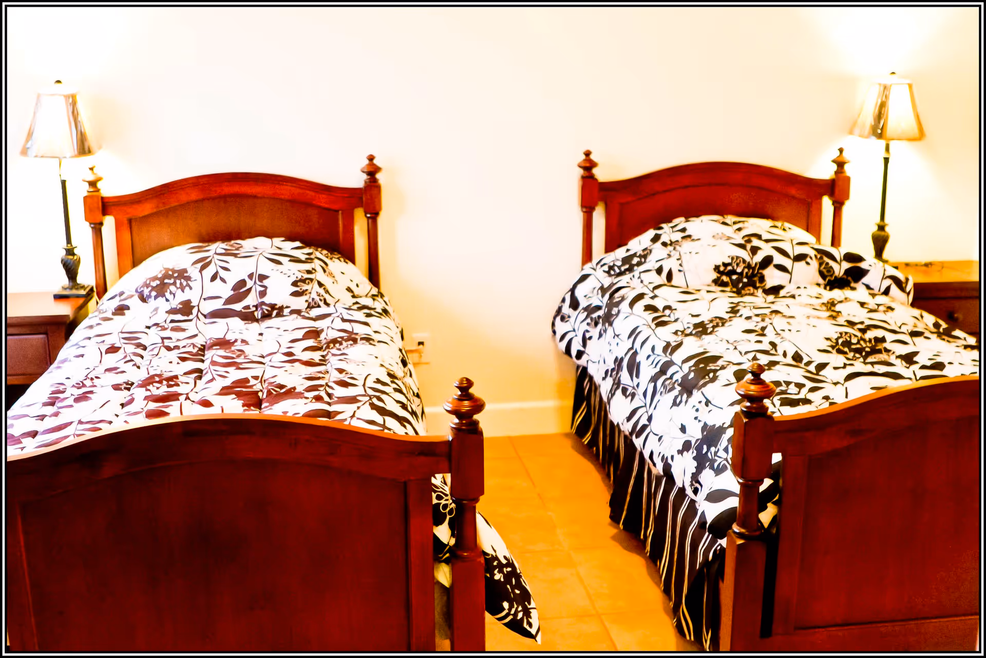 A bedroom with two wooden twin beds, each with a floral patterned comforter in black and white. There are two bedside tables, each with a lamp, against a plain white wall.