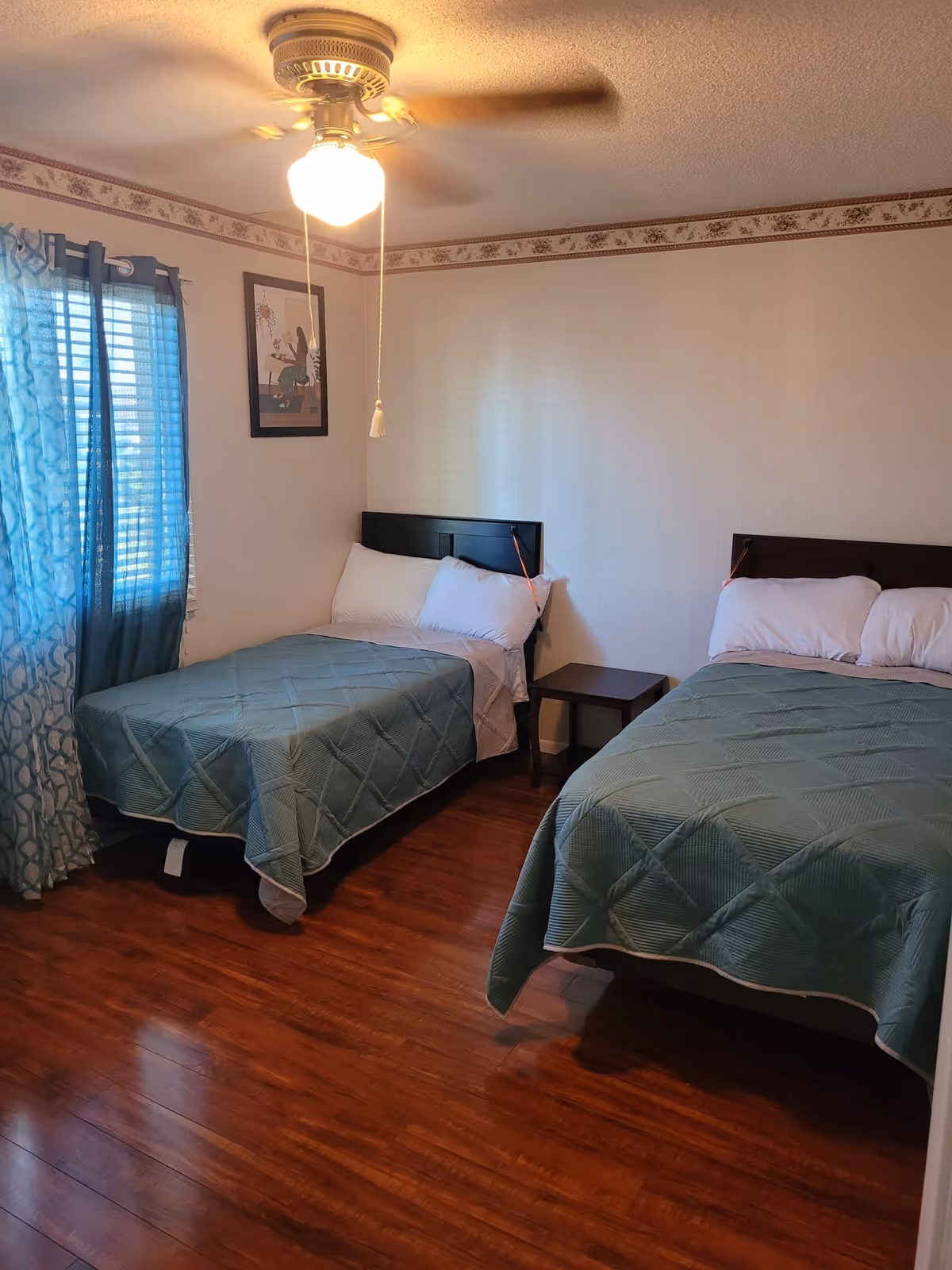 A bedroom with two beds covered in green quilts, each with white pillows. There is a small wooden nightstand between the beds. A ceiling fan with a light is on, and a window with blue patterned curtains is on the left wall. A framed picture hangs on the wall above the left bed. The floor is wooden, and the walls are light-colored with a floral border near the ceiling.