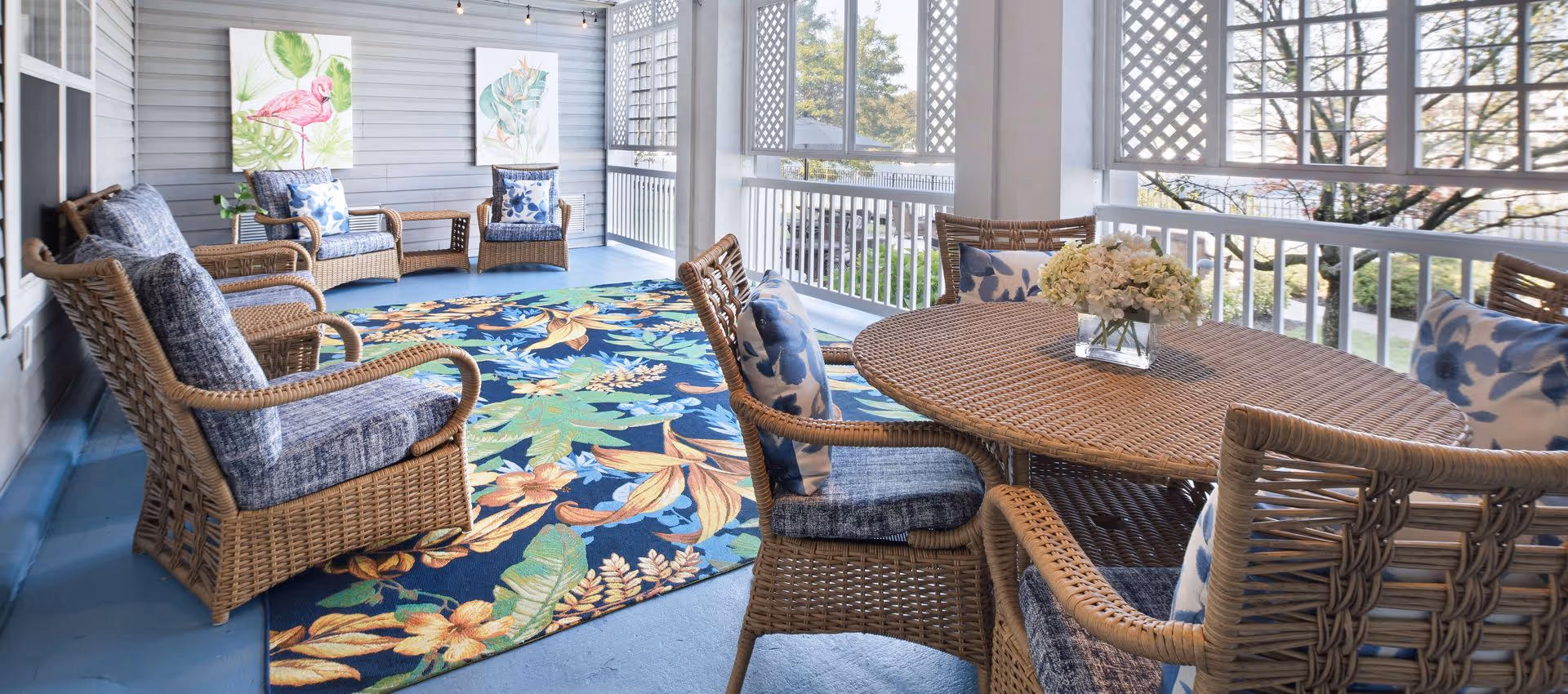 Covered porch with wicker chairs and a round dining table on a colorful floral rug overlooking a garden.