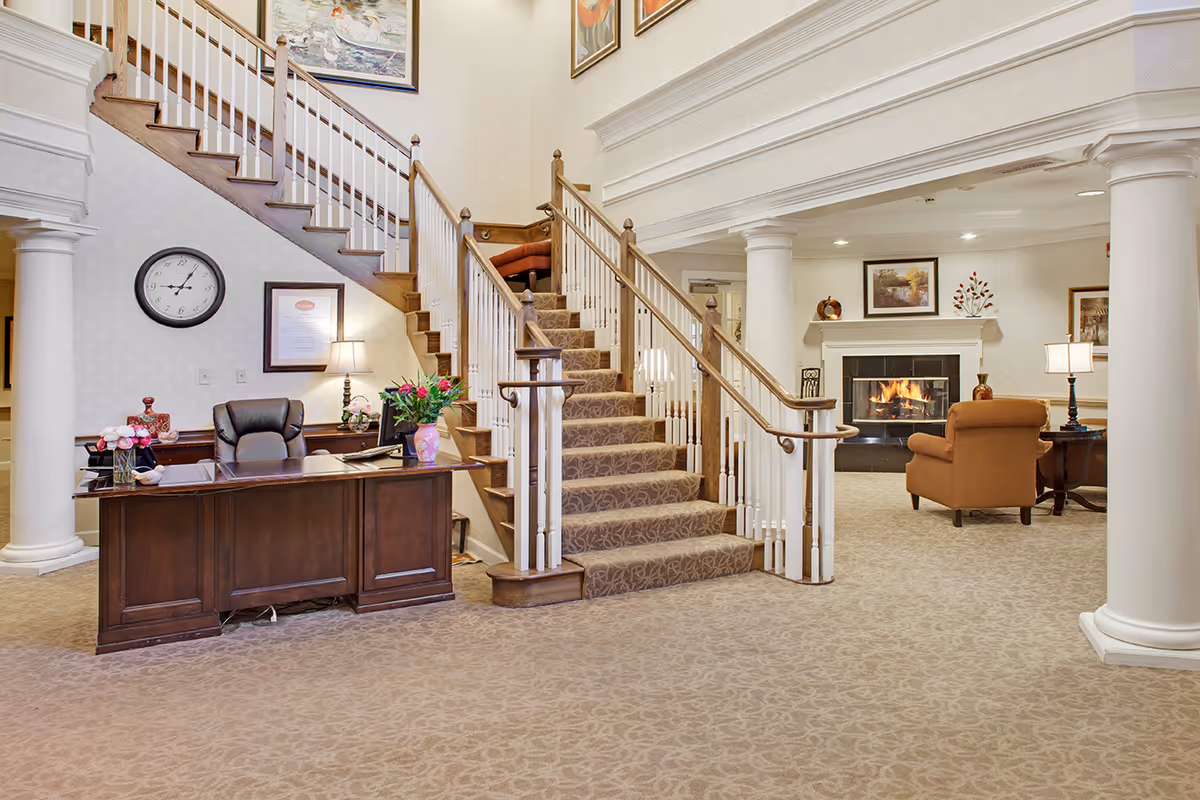 Interior view of a senior living facility lobby with a wooden reception desk, a staircase with carpeted steps and white railings, a cozy seating area with an armchair near a fireplace, and decorative columns.
