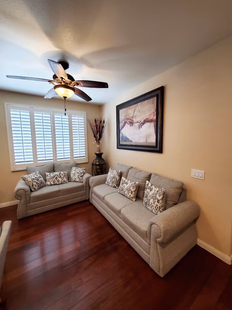 Cozy living room with two gray sofas and patterned pillows, a ceiling fan, shuttered window, wall art, and hardwood floors.