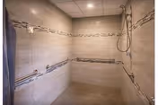 Spacious walk-in shower with beige tiled walls and floor, featuring decorative horizontal tile accents. The shower has a handheld showerhead, grab bars on two walls, and a built-in bench seat.