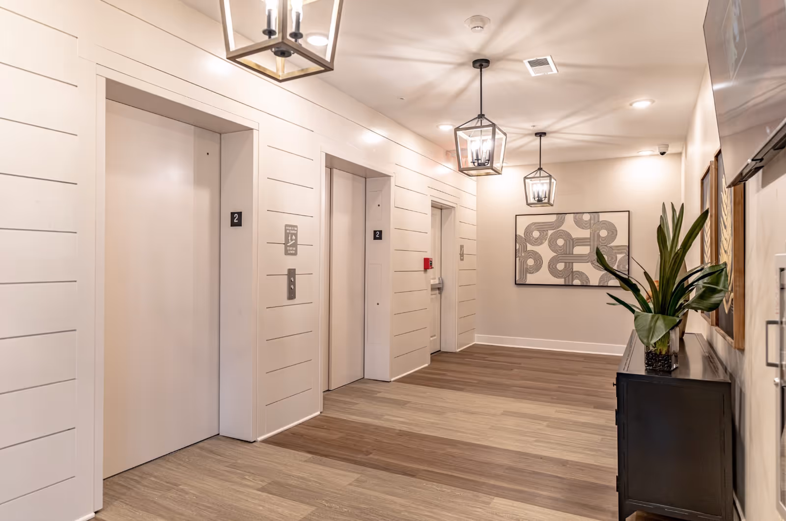 Interior hallway with three elevator doors labeled with the number 2, modern pendant lights hanging from the ceiling, a decorative plant on a black console table, and abstract artwork on the walls.