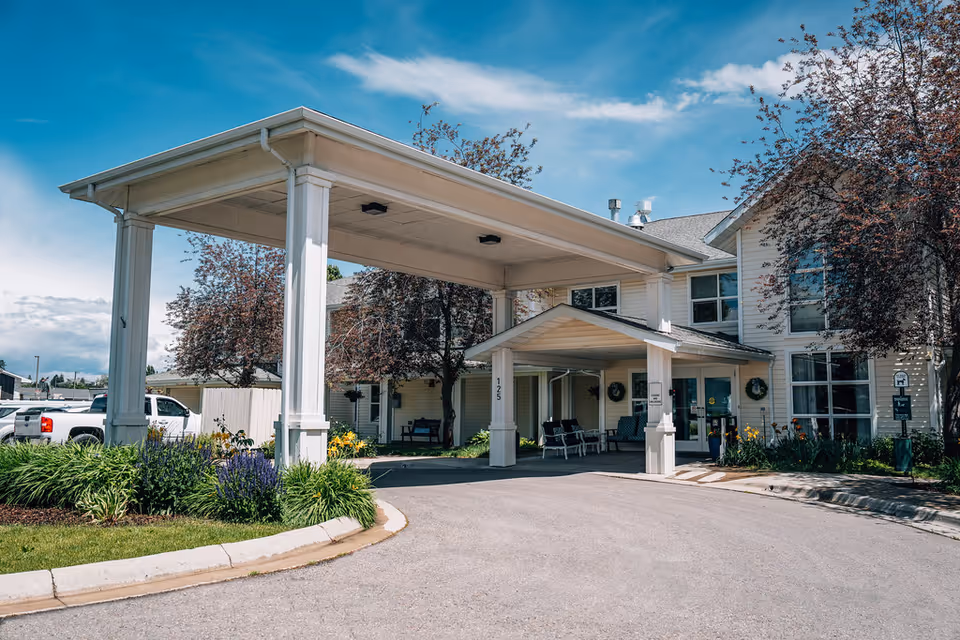 Exterior view of Kalispell Assisted Living facility showing the main entrance with a covered drop-off area, surrounded by landscaped greenery and trees under a blue sky.