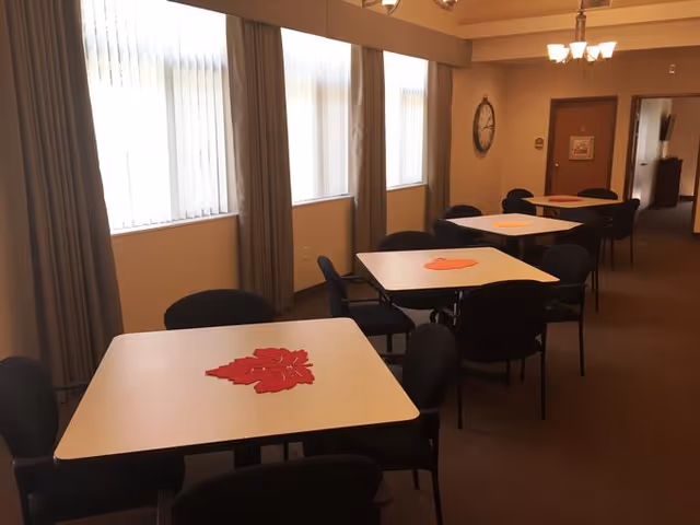Interior view of a dining area with several square tables, each decorated with a red or orange leaf-shaped centerpiece. Black chairs surround the tables. Large windows with vertical blinds and curtains allow natural light into the room. A wall clock and a chandelier light fixture are visible on the beige walls.
