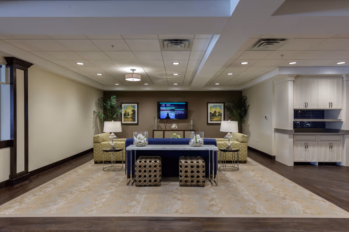 A spacious and well-lit living room area in Clarendale of Algonquin featuring a large beige patterned rug, a blue sofa with two patterned ottomans underneath a console table, two yellow armchairs with side tables and lamps, two framed landscape paintings on a brown accent wall, a flat-screen TV mounted on the wall, and built-in cabinetry with a dark blue tile backsplash on the right side.
