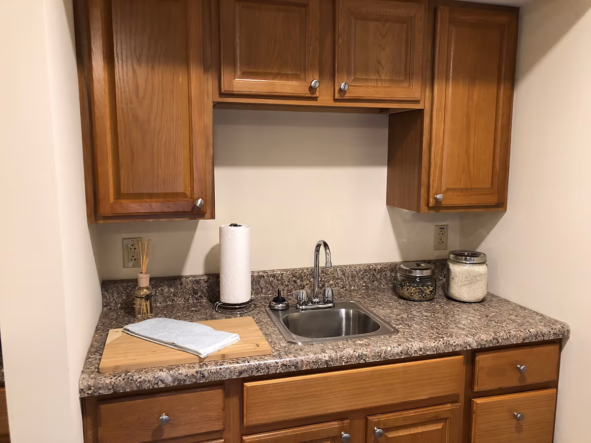 A small kitchen area with wooden cabinets, a granite countertop, and a stainless steel sink. On the countertop, there is a paper towel roll, a wooden cutting board with a cloth on it, a reed diffuser, and two glass jars containing dry goods.