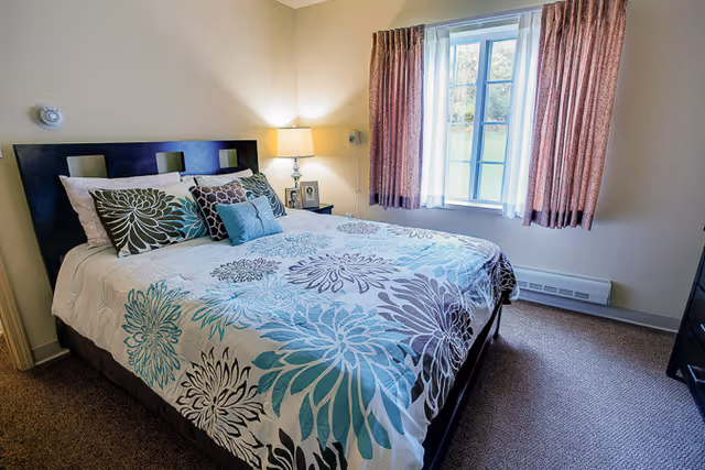 A neatly made bed with a floral patterned bedspread in shades of blue, teal, and gray in a bedroom. The bed has multiple pillows, including decorative ones. A bedside table with a lamp and framed photos is next to the bed. A window with pinkish curtains lets in natural light, illuminating the beige walls and carpeted floor.