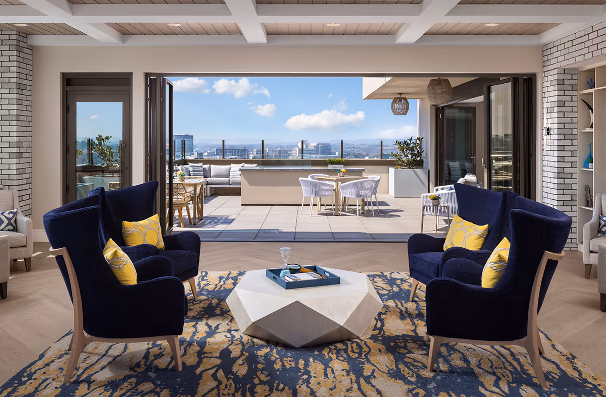 A modern living room with four navy blue armchairs featuring yellow patterned pillows arranged around a geometric white coffee table on a blue and beige patterned rug. The room opens up to a spacious outdoor patio with a seating area, dining table, and a view of the city skyline under a partly cloudy sky.