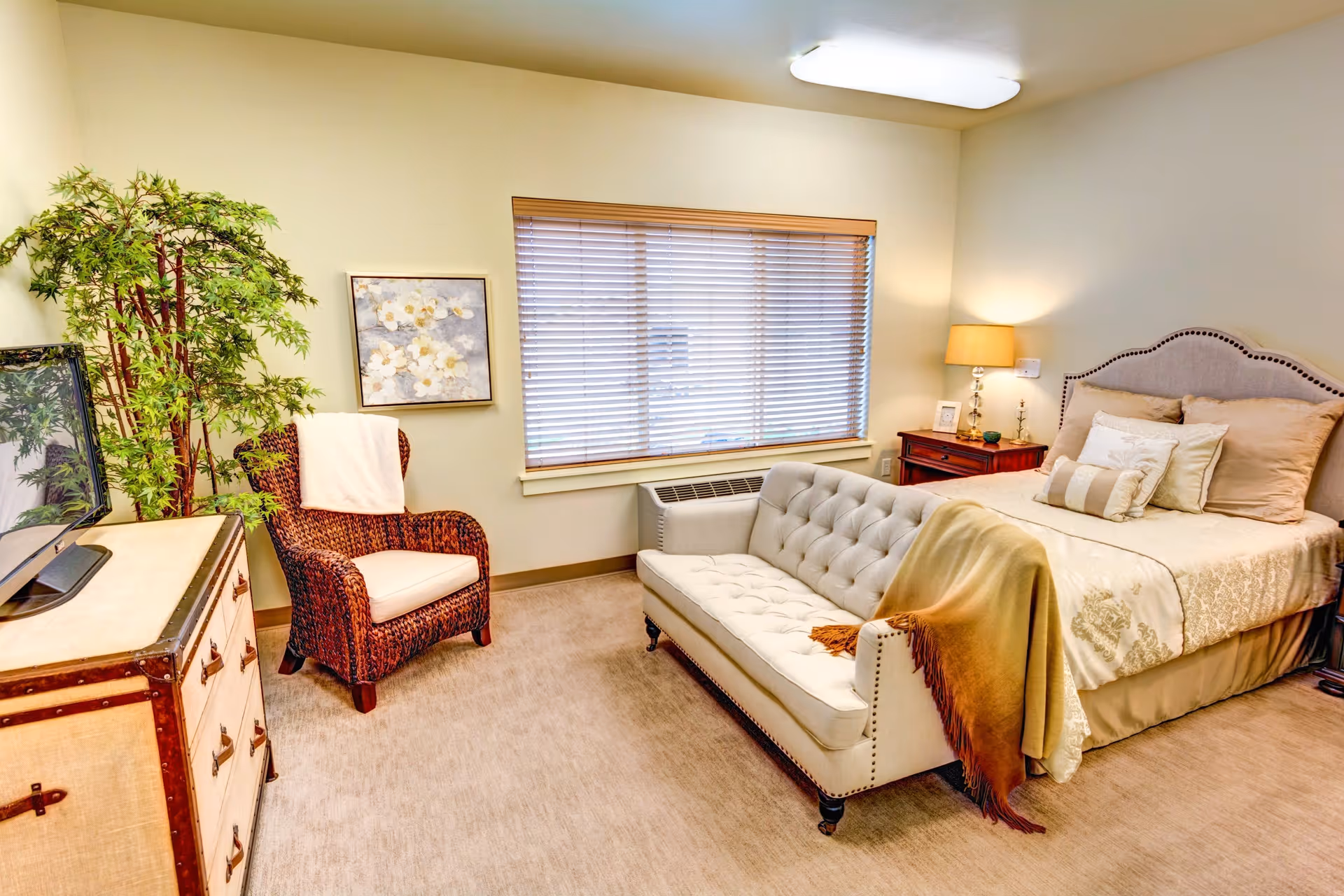 A cozy bedroom in a senior living facility featuring a large bed with multiple pillows and a beige bedspread. At the foot of the bed is a tufted cream-colored loveseat with a brown and beige throw blanket draped over one side. To the left, there is a wicker armchair with a white cushion and a white throw blanket. A wooden dresser with a TV on top is positioned against the wall, and a large potted plant stands beside it. The room has beige carpet, a window with wooden blinds, a nightstand with a lamp, and a framed floral painting on the wall.