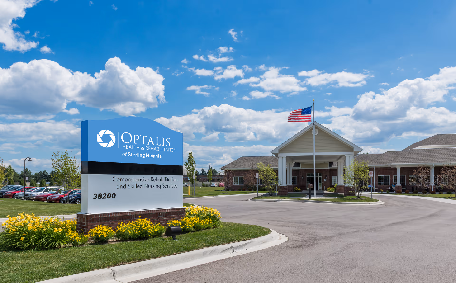 Exterior view of Optalis Health & Rehabilitation of Sterling Heights facility with a large sign in the foreground displaying the facility name and services. The building has a covered entrance with an American flag flying on a flagpole. The sky is blue with scattered clouds, and there are parked cars and landscaped greenery around the area.