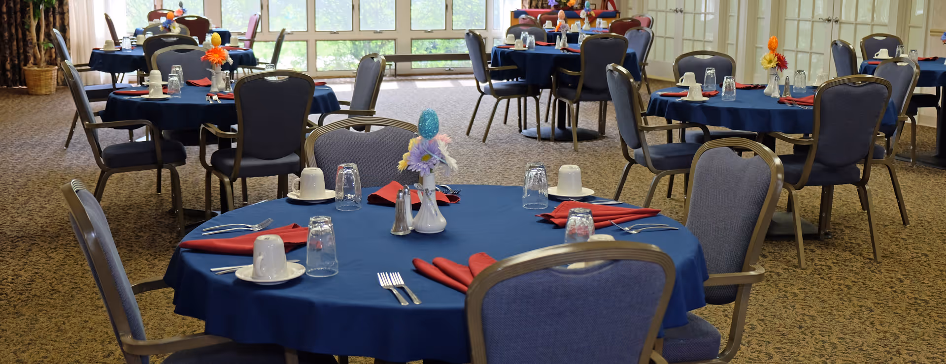 A dining room with multiple round tables covered with blue tablecloths, each set with white cups on saucers, upside-down glasses, silverware, red folded napkins, and small floral centerpieces. The room has carpeted flooring and large windows letting in natural light.