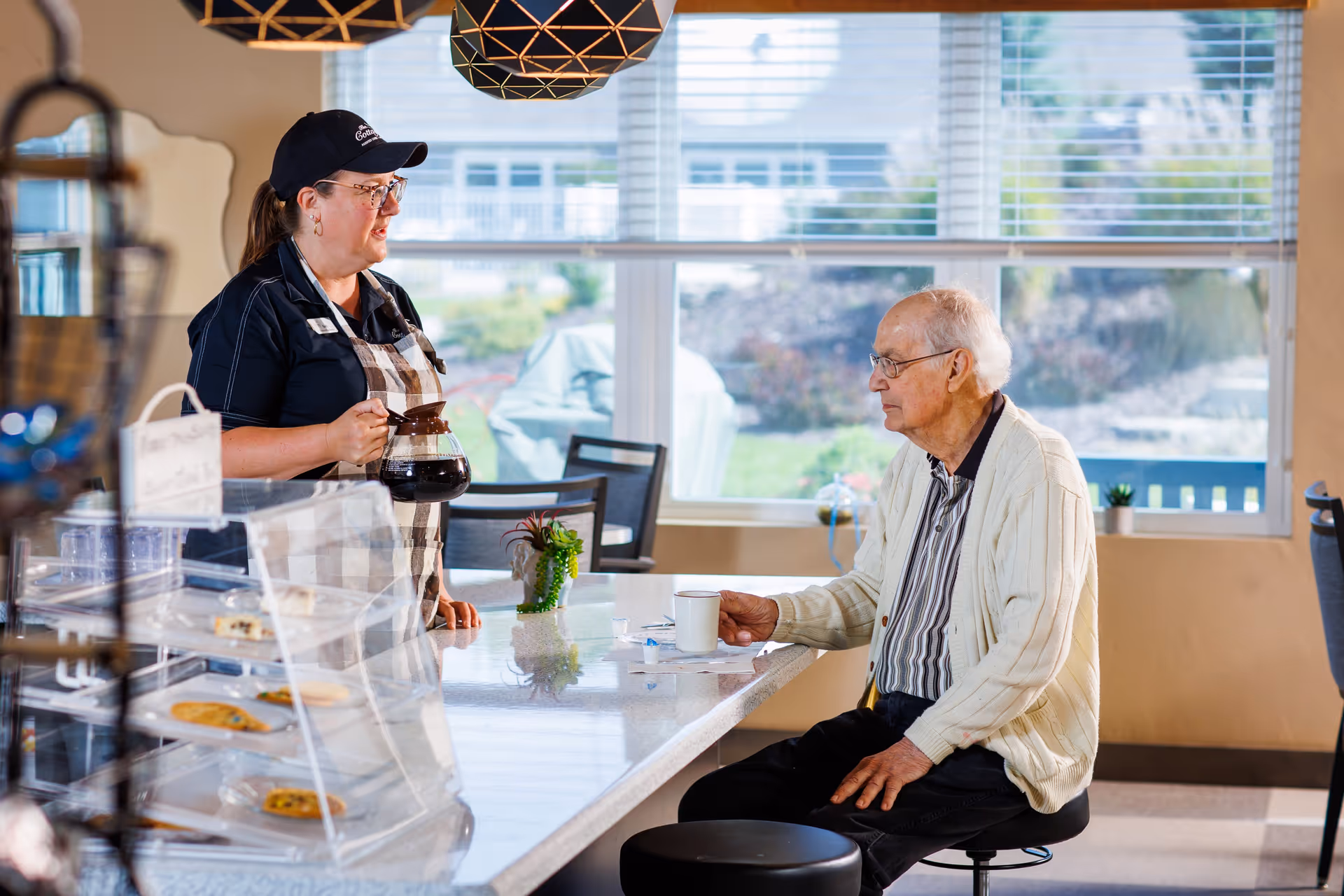 A senior man sitting on a stool at a counter in a bright room with large windows. A woman wearing a black cap and apron is standing behind the counter, holding a coffee pot and talking to the man. There are some cookies displayed in a clear case on the counter and a small plant decoration.
