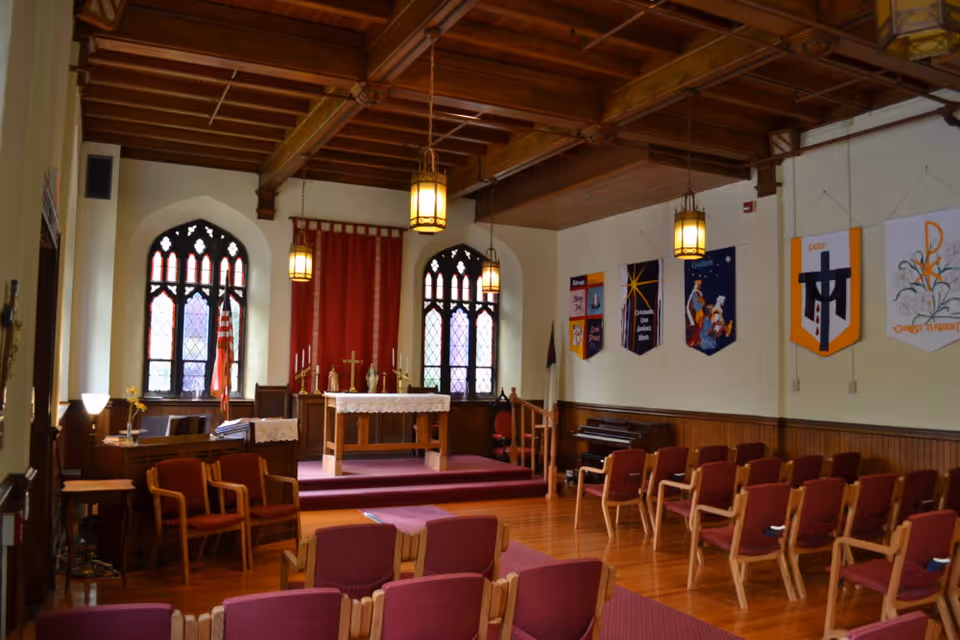 Interior of a chapel or small worship room with wooden ceiling beams, stained glass windows, hanging lantern lights, an altar with candles and a cross, rows of wooden chairs with red cushions, and religious banners hanging on the wall.