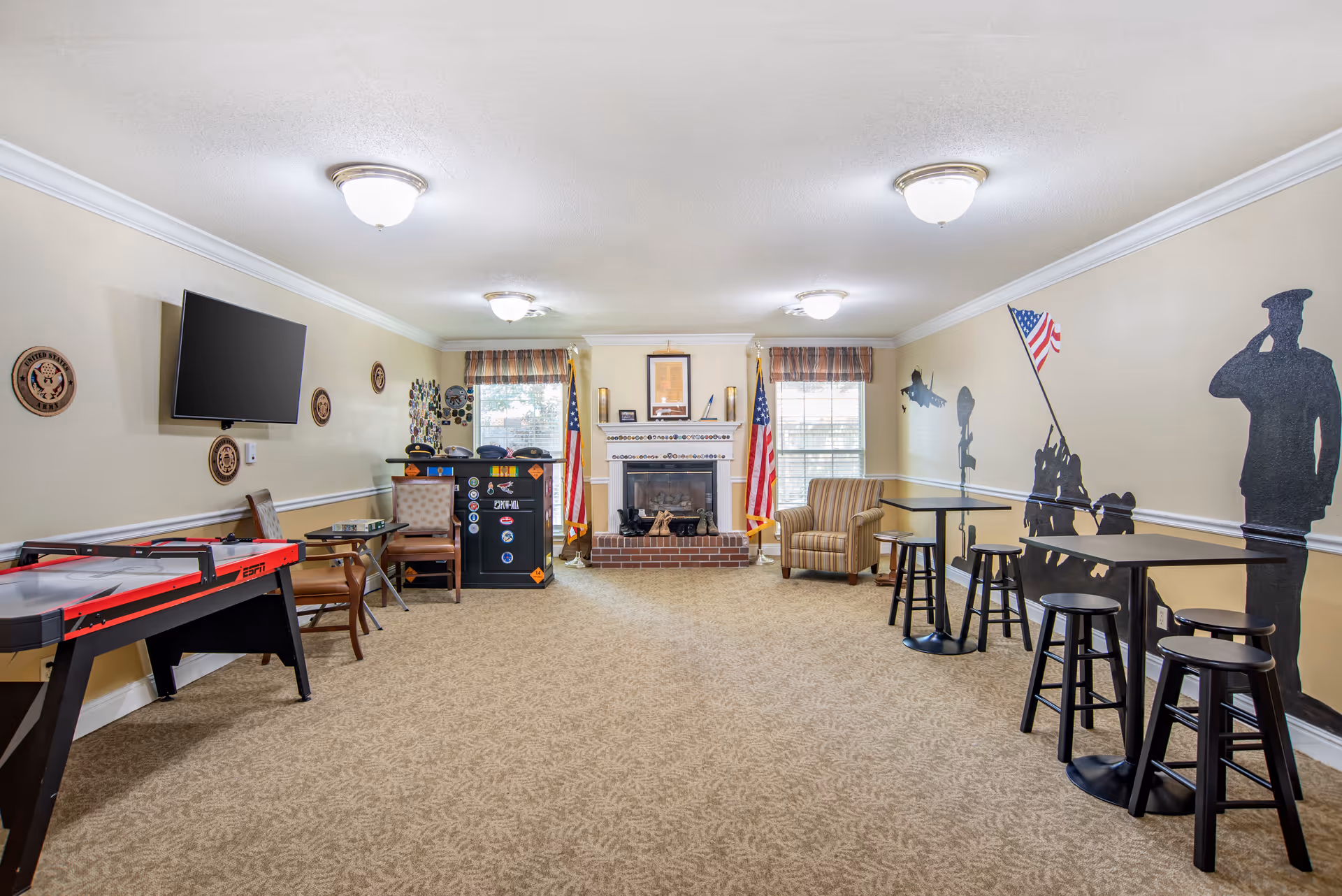 A spacious common room with beige carpet and cream walls featuring military-themed decorations including wall plaques, a mural of soldiers saluting and raising an American flag, and two American flags flanking a brick fireplace. The room has a mounted flat-screen TV, several chairs, small tables with stools, and an air hockey table on the left side.