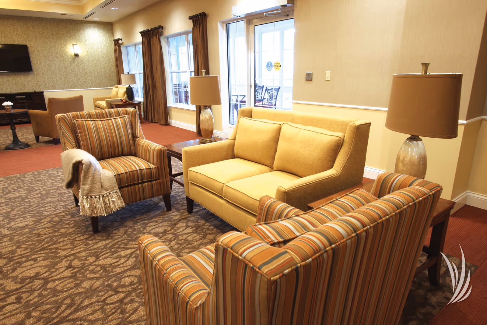 Comfortable senior living room with a yellow loveseat, striped armchairs, side tables and lamps near large windows and glass doors.