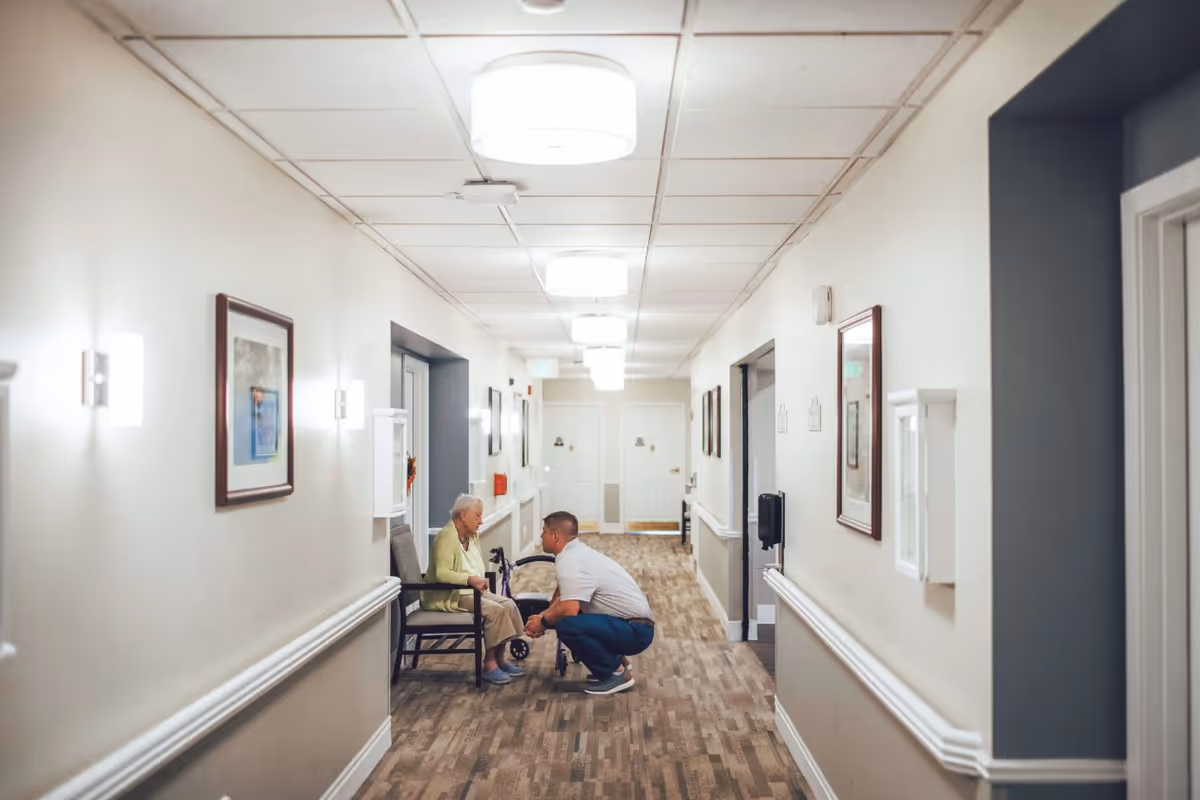 A hallway in an assisted living facility with beige walls and carpeted floor. An elderly woman is seated in a chair along the wall, and a man is crouching down in front of her, engaging in conversation. The hallway is well-lit with ceiling lights and wall sconces, and framed pictures hang on the walls.