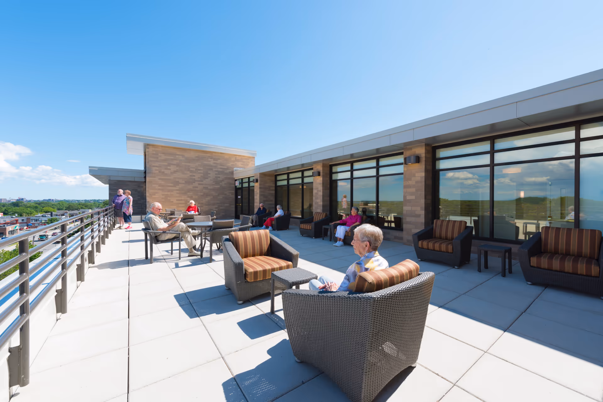 A rooftop patio at The Park Danforth with several elderly people sitting on wicker chairs with striped cushions and small tables. The patio has a railing on one side and large windows on the building side, under a clear blue sky.