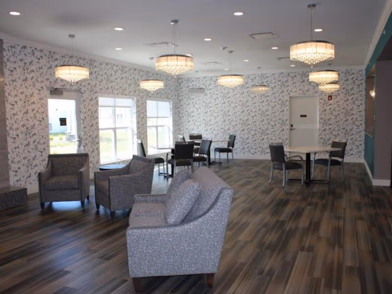 A spacious, well-lit common area with floral wallpaper, multiple round tables with chairs, and several cushioned armchairs and a sofa. The room has large windows letting in natural light and modern ceiling light fixtures.