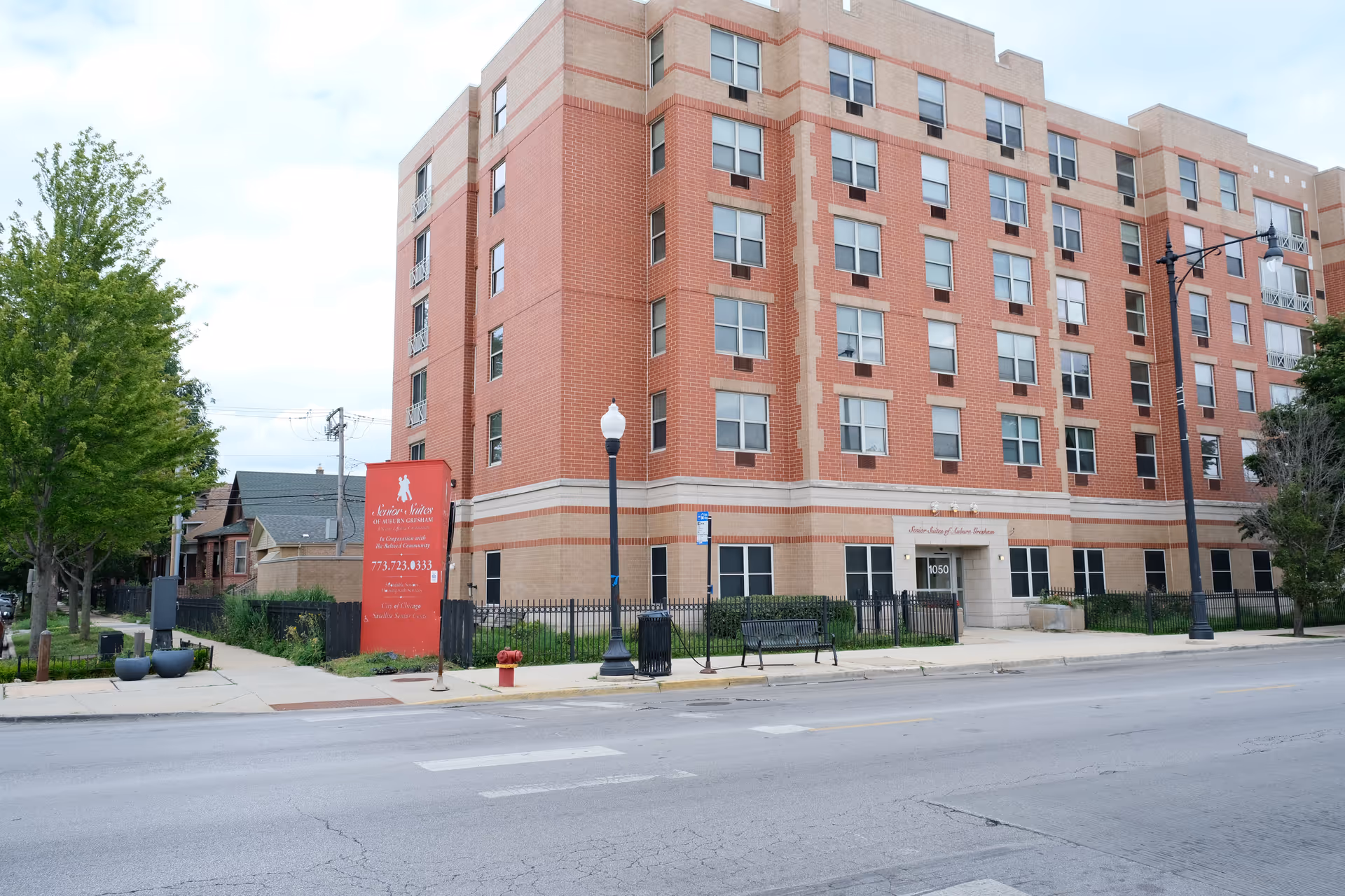 Front view of a multi-story brick senior living building with a red sign and sidewalk in front.