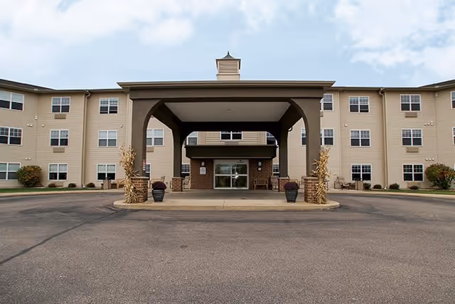 Front exterior view of a three-story senior living facility with a covered entrance and a circular driveway. The building has beige siding, multiple windows, and decorative plants near the entrance.