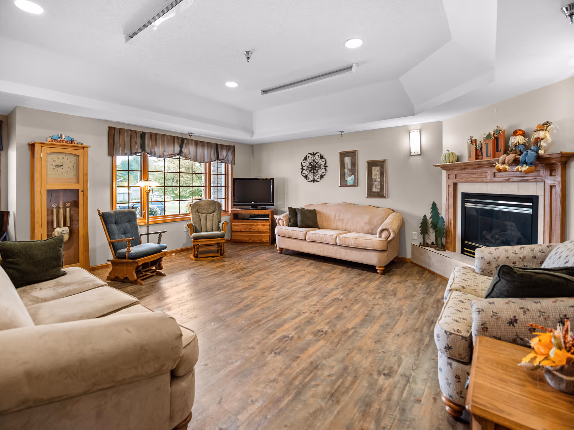 A cozy living room with wooden flooring, beige and patterned sofas, two wooden rocking chairs, a wooden grandfather clock, a TV on a wooden stand, and a fireplace decorated with autumn-themed items. A large window with brown valances lets in natural light.
