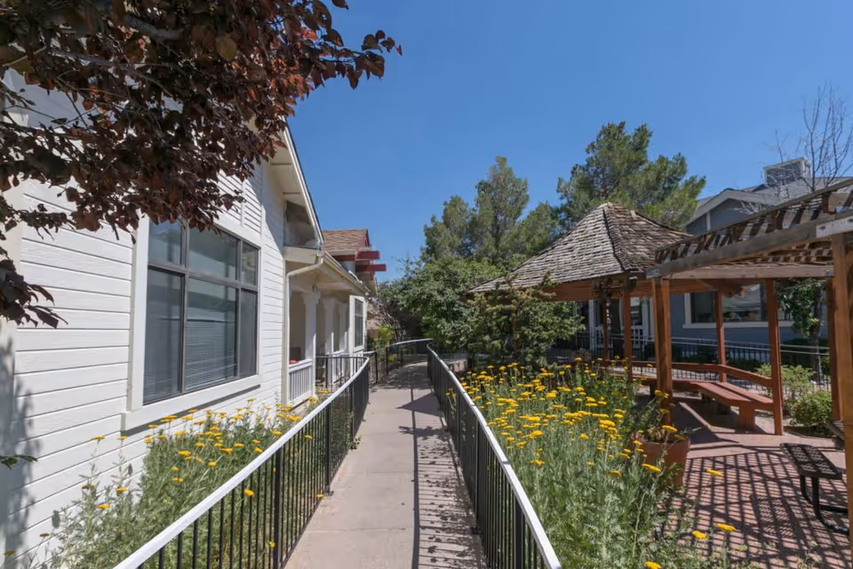 Outdoor walkway at Park Place Assisted Living with a white building on the left, a black metal railing along the path, yellow flowers growing beside the walkway, and a wooden gazebo with benches on the right under a clear blue sky.
