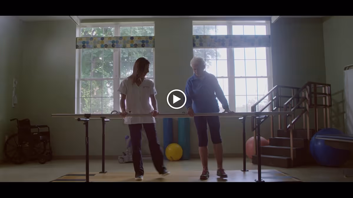 A physical therapy room with two women standing and holding parallel bars. One woman appears to be a healthcare professional assisting an elderly woman with walking exercises. The room has large windows with patterned valances, exercise balls, a wheelchair, and a small staircase with handrails.