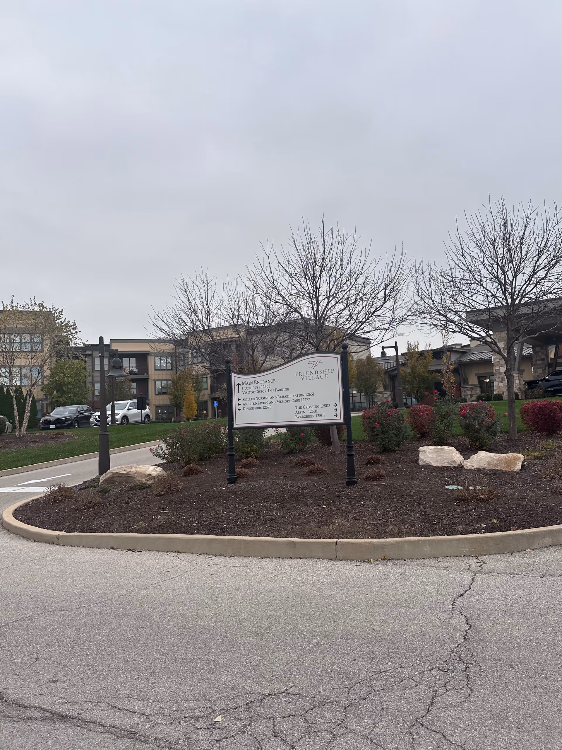 Outdoor view of the entrance area at Friendship Village Sunset Hills, featuring a directional sign surrounded by landscaping with bushes and leafless trees, with buildings and parked cars in the background under a cloudy sky.