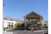 Exterior view of Millard County Care Center building under a clear blue sky, featuring a covered entrance with a peaked roof, landscaped area with plants and a circular driveway in front.