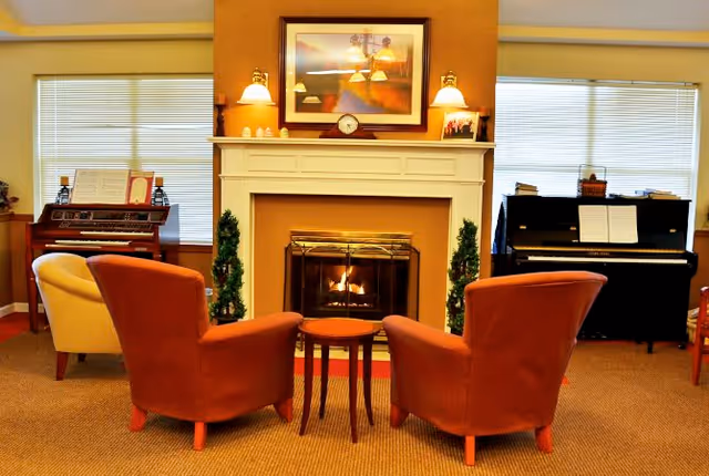 A cozy living room area with a lit fireplace in the center, flanked by two small green plants. In front of the fireplace are two orange armchairs and a small round wooden table. On either side of the fireplace are two pianos, one upright and one organ-style, with sheet music on them. The walls are painted a warm yellow, and there are two windows with blinds on each side of the room.