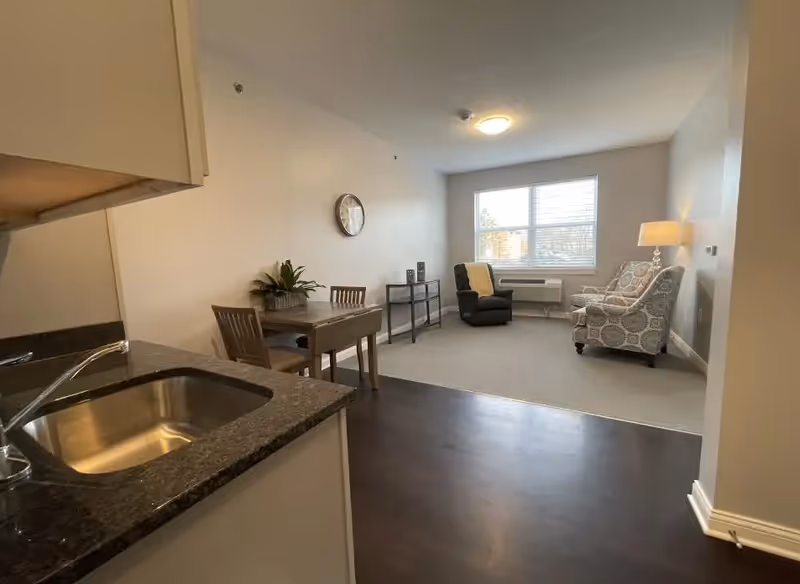 Interior view of a living space in Autumn Trace Bedford Assisted Living showing a small kitchen area with a sink and countertop in the foreground, a wooden dining table with two chairs, and a living area with two armchairs, a side table with a lamp, a window with blinds, and a wall clock.