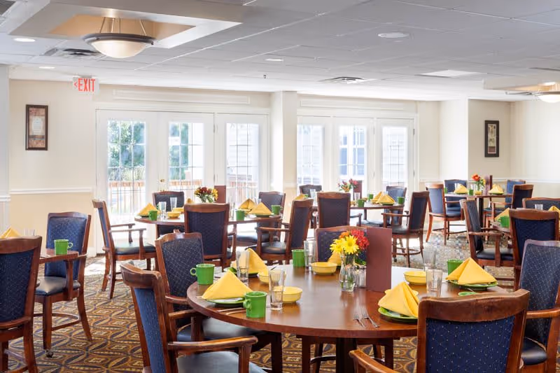 A bright dining room with multiple round wooden tables set with yellow napkins, green mugs, glasses, and silverware. Each table has a small vase with colorful flowers. The room has large windows and glass doors letting in natural light, blue cushioned wooden chairs, and patterned carpet flooring.