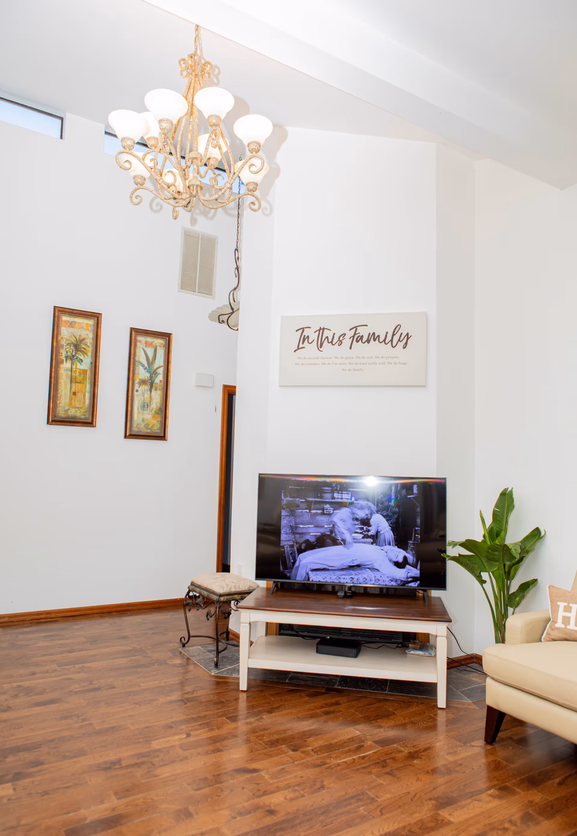 A bright living room with wooden flooring, a beige armchair with a pillow, a flat-screen TV on a wooden stand, a green potted plant, two framed pictures on the wall, and a decorative chandelier hanging from the ceiling. Above the TV, there is a wall sign that reads 'In this Family'.
