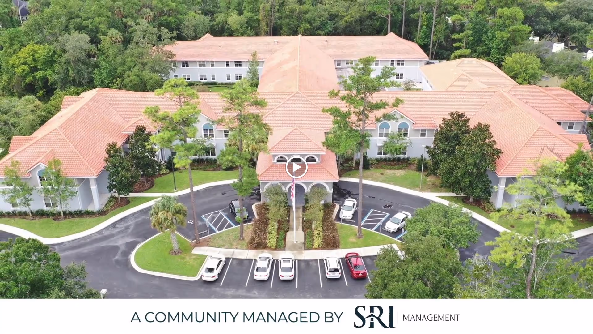Aerial view of The Cove at Marsh Landing senior living facility showing a large building with a red-tiled roof surrounded by trees and greenery. The building has a circular driveway with several parked cars in front and landscaped areas with palm trees and bushes.
