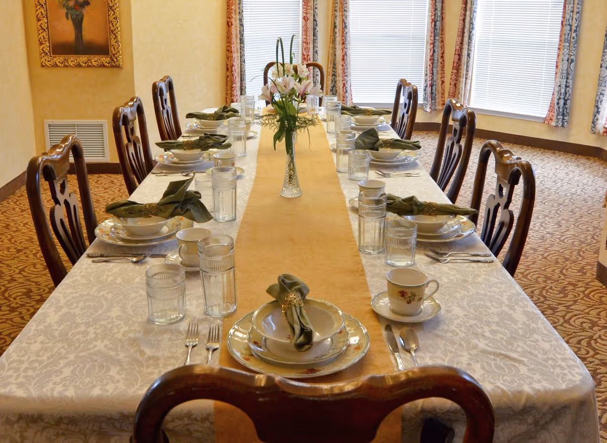 A long dining table set for a meal with white tablecloth and a beige table runner. The table is arranged with plates, bowls, cups, glasses, silverware, and green cloth napkins with napkin rings. A vase with flowers is placed in the center of the table. The room has patterned carpet, beige walls, and windows with blinds and floral curtains.