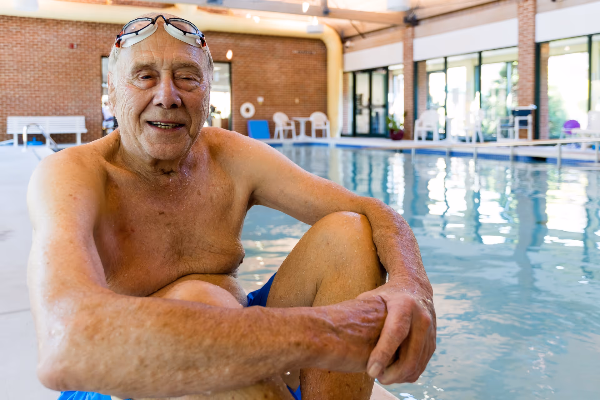 An elderly man wearing swimming goggles on his forehead sits at the edge of an indoor swimming pool, smiling. The background shows a spacious pool area with large windows, brick walls, and poolside chairs.