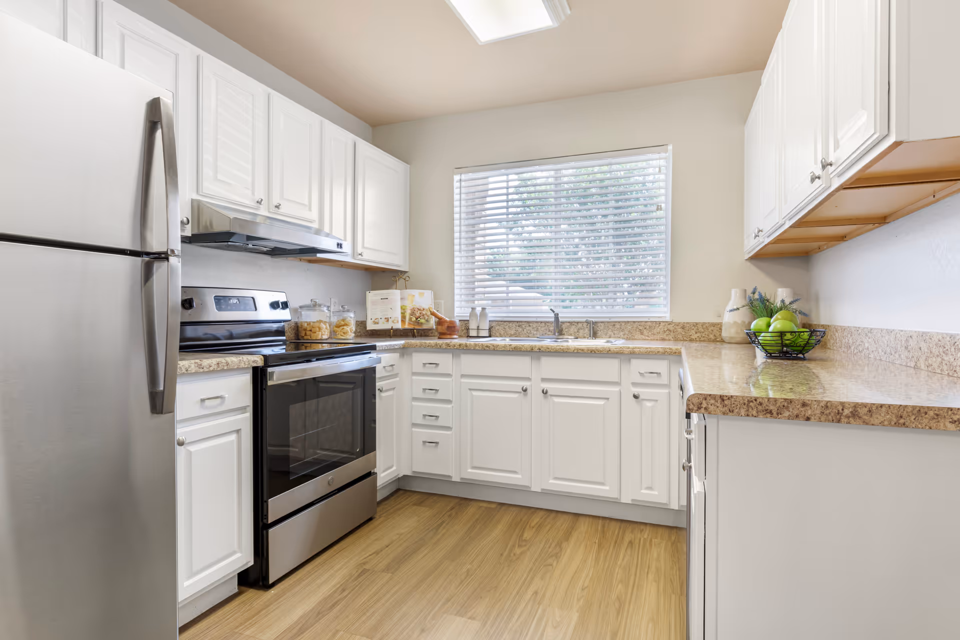 Bright kitchen with white cabinets, stainless steel refrigerator and oven, beige granite countertops, wooden floor, and a window with blinds letting in natural light. The countertop has jars, a cookbook, a mortar and pestle, decorative vases, and a bowl of green apples.
