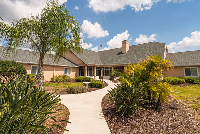 Front exterior of a single-story assisted living building with a curved walkway, palm trees, and landscaped beds under a partly cloudy sky.