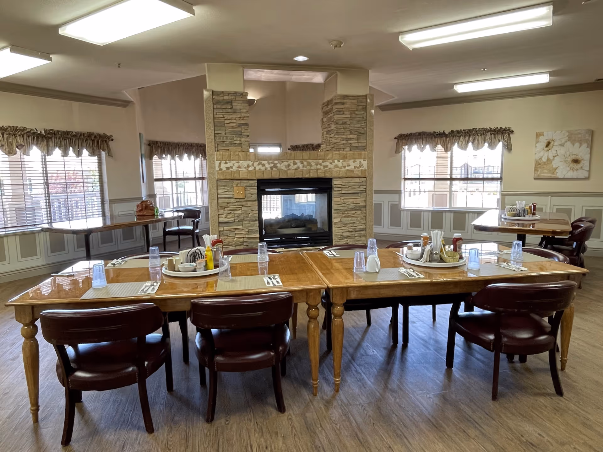 A dining room in an assisted living facility with wooden tables and chairs arranged around a central stone fireplace. The tables are set with placemats, plastic cups, and condiment trays. Large windows with valance curtains allow natural light to fill the room, and a floral painting hangs on the wall.