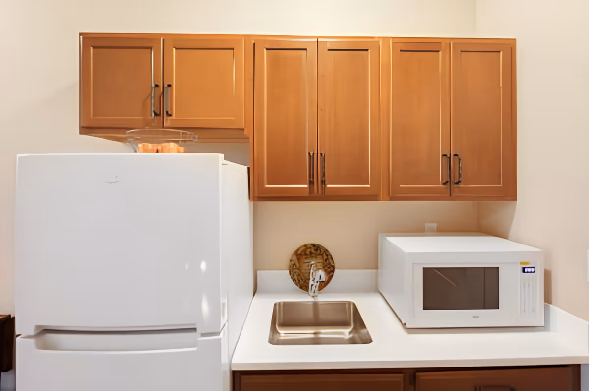 A small kitchen area with wooden cabinets mounted on the wall, a white refrigerator on the left, a stainless steel sink in the center of the countertop, and a white microwave on the right side of the countertop.