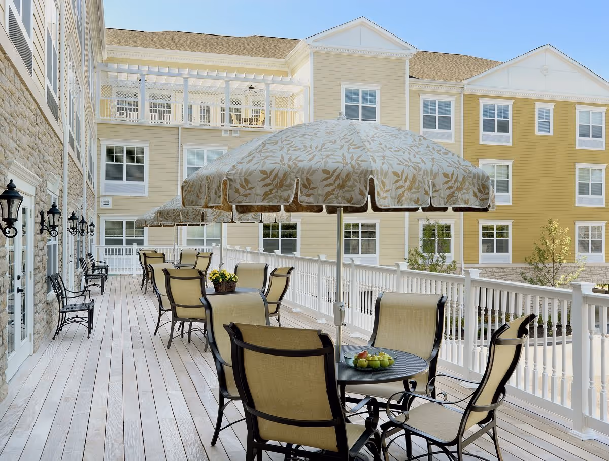 Outdoor patio area with multiple tables and chairs under large patterned umbrellas. The patio has a wooden floor and is adjacent to a multi-story building with beige and yellow siding and white railings.