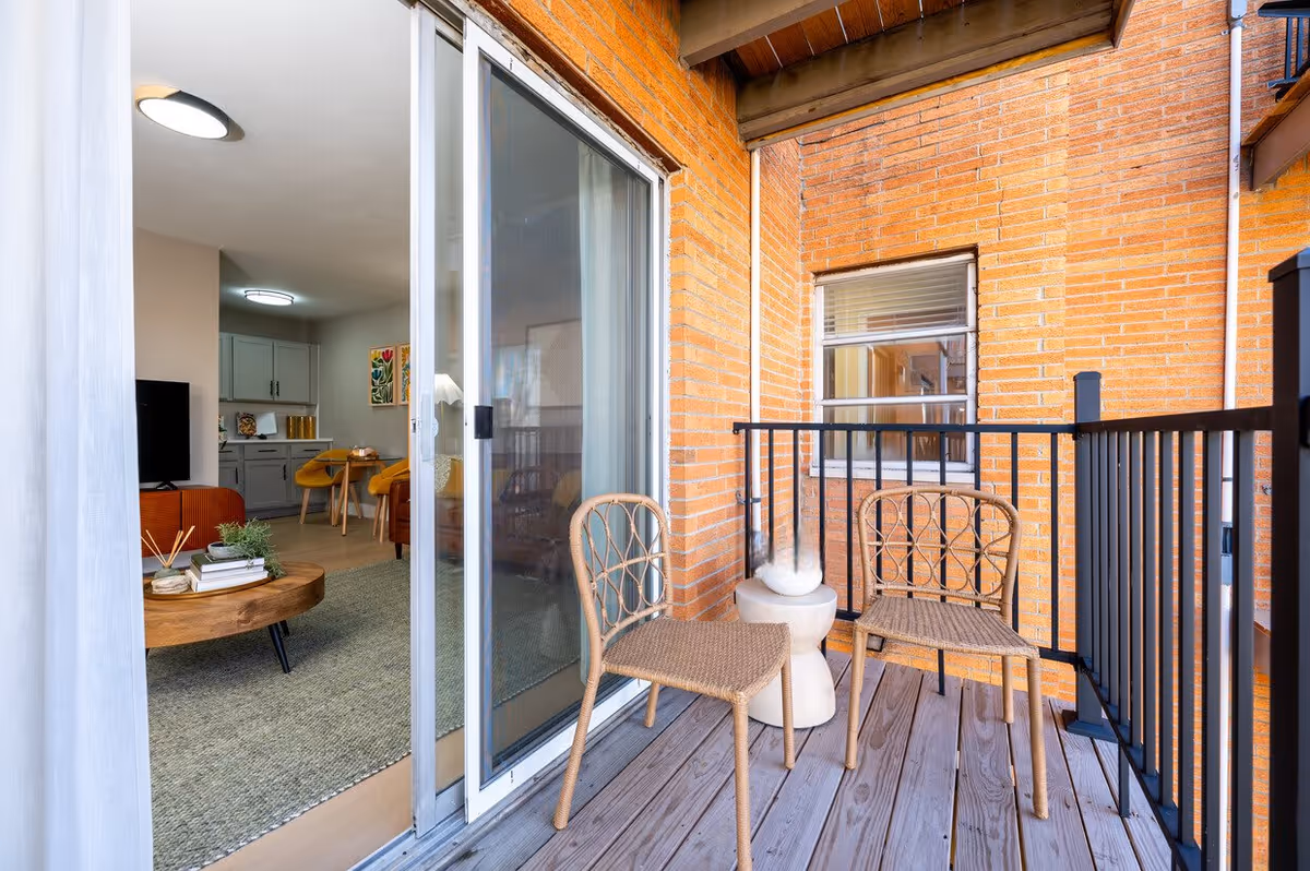 A small balcony with two wicker chairs and a small white side table. The balcony has wooden flooring and black metal railing. The balcony is adjacent to a brick wall and a window. Through a sliding glass door, an interior living space is visible with a round wooden coffee table, a TV, and a dining area with yellow chairs and gray cabinets.