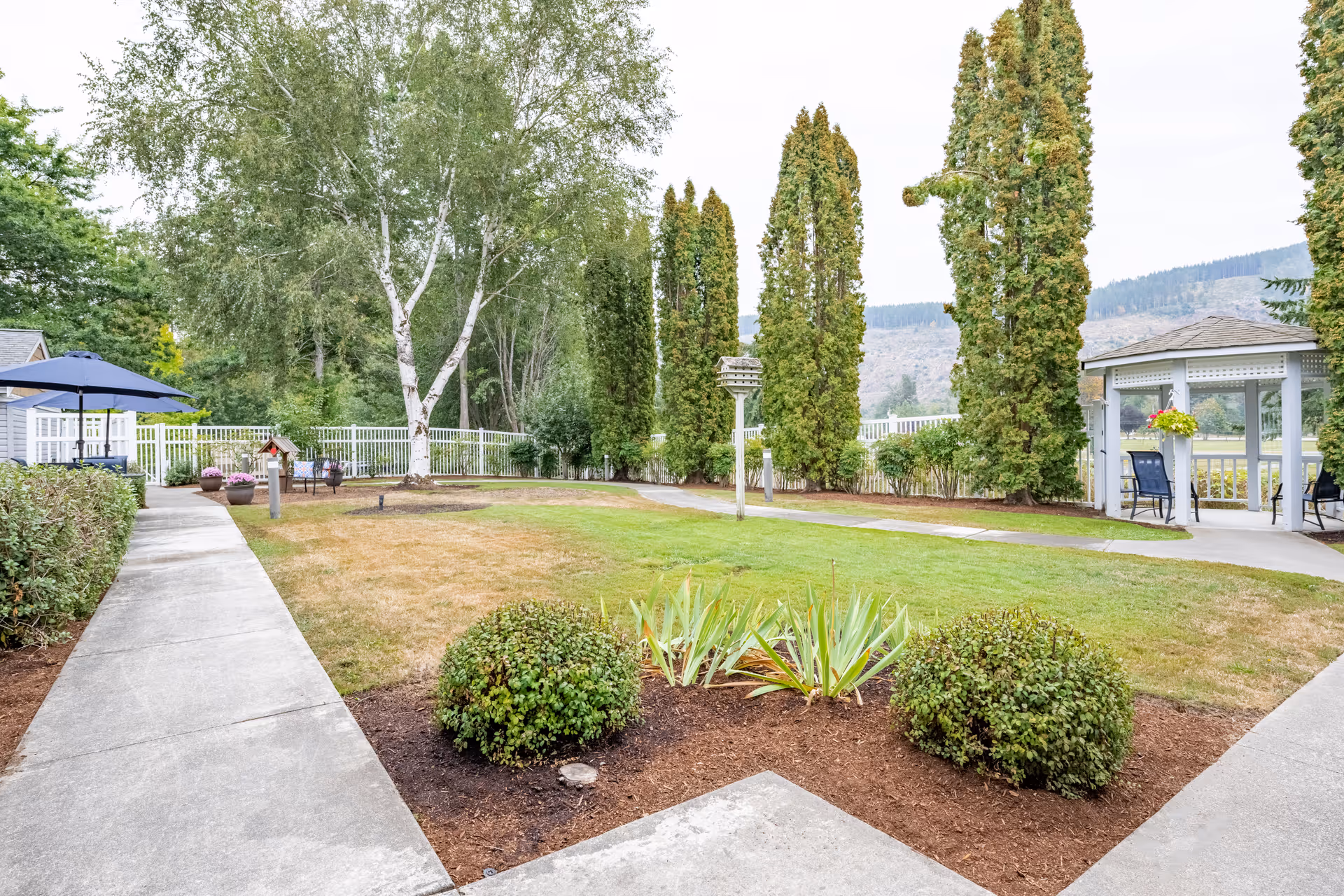 A well-maintained outdoor garden area at Birchview Memory Care featuring a concrete pathway, green grass, trimmed bushes, tall trees, a birdhouse, and a white gazebo with seating. The background includes a white fence and distant hills.