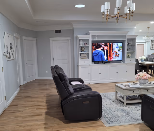 A cozy living room area with two black recliner chairs facing a white entertainment center with a flat-screen TV displaying a scene with two people. The room has light wood flooring, a white coffee table with a flower arrangement, and a chandelier hanging from the ceiling. In the background, there is a dining area with a table and chairs.