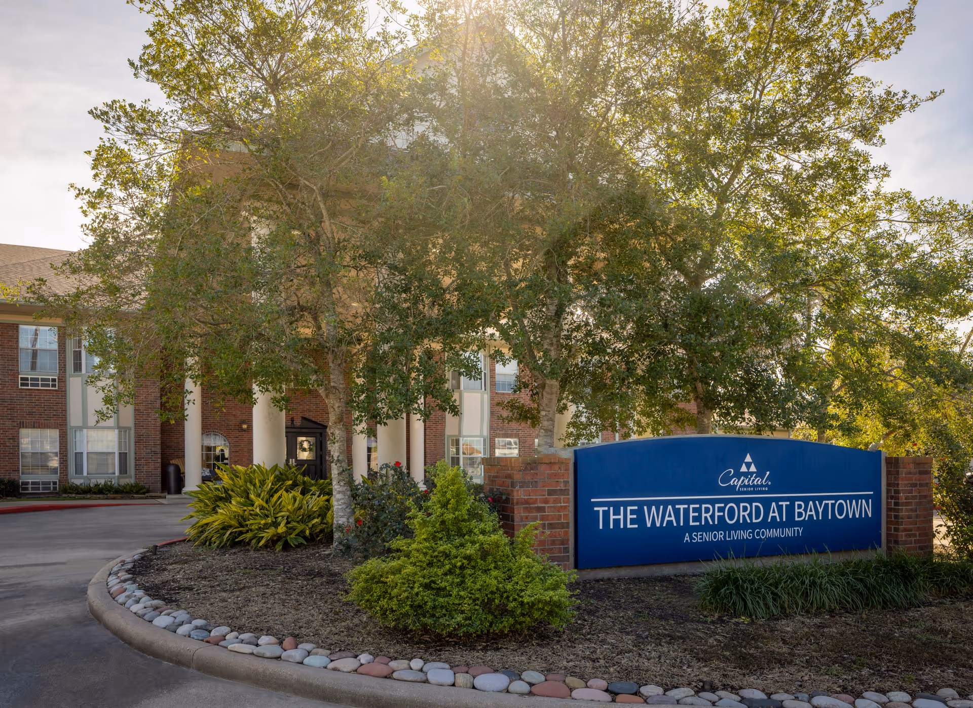 Exterior view of The Waterford at Baytown senior living community building with a large blue sign in front surrounded by trees and landscaping under a sunny sky.