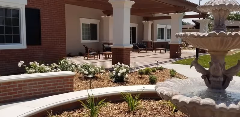 Outdoor patio area of a senior living facility with a multi-tiered water fountain in the foreground, surrounded by landscaping with plants and mulch. The building features brick and white columns supporting a covered seating area with chairs and benches.
